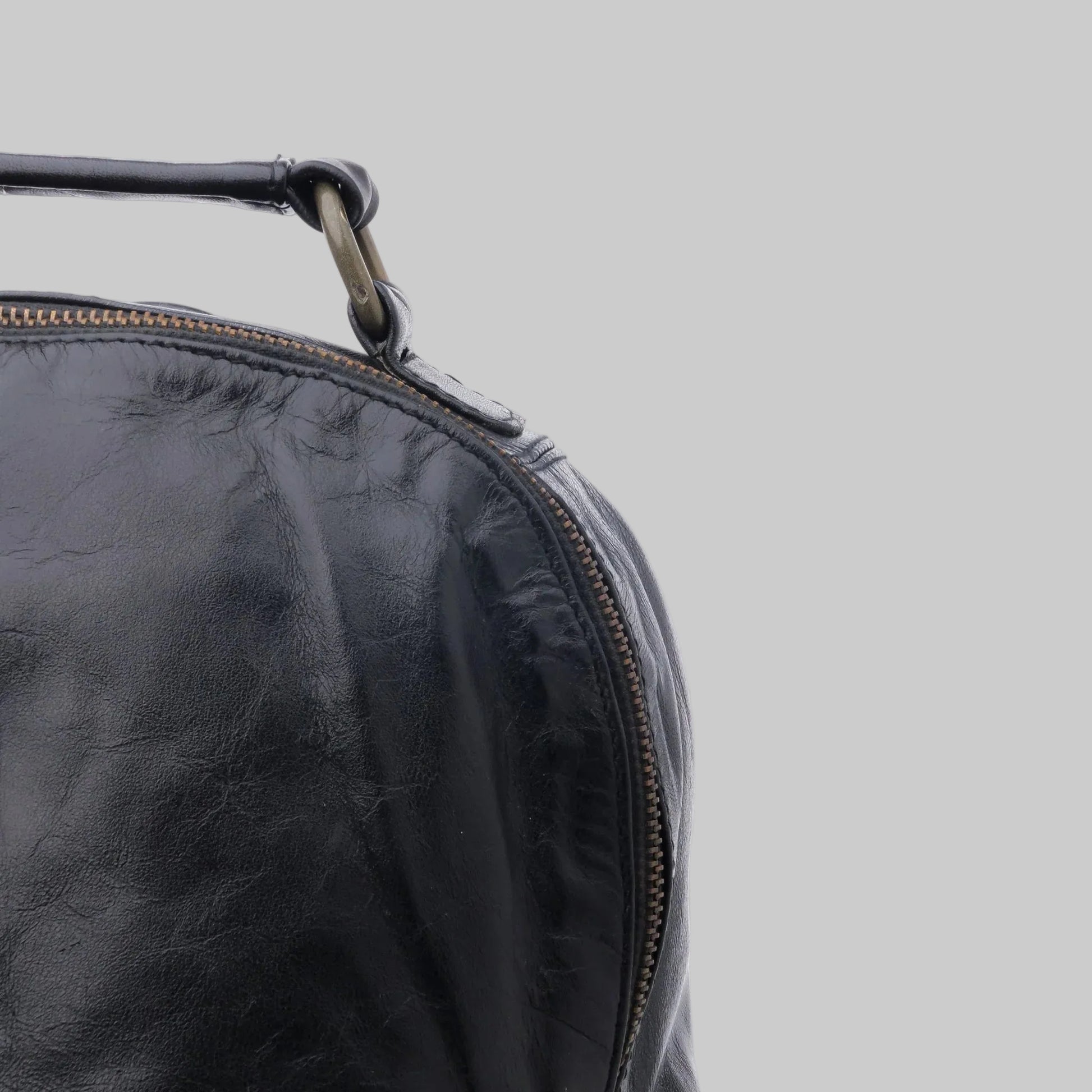 Close-up of a black leather bag corner showing a curved zipper and a dark strap attached with a metal ring, against a plain white background.