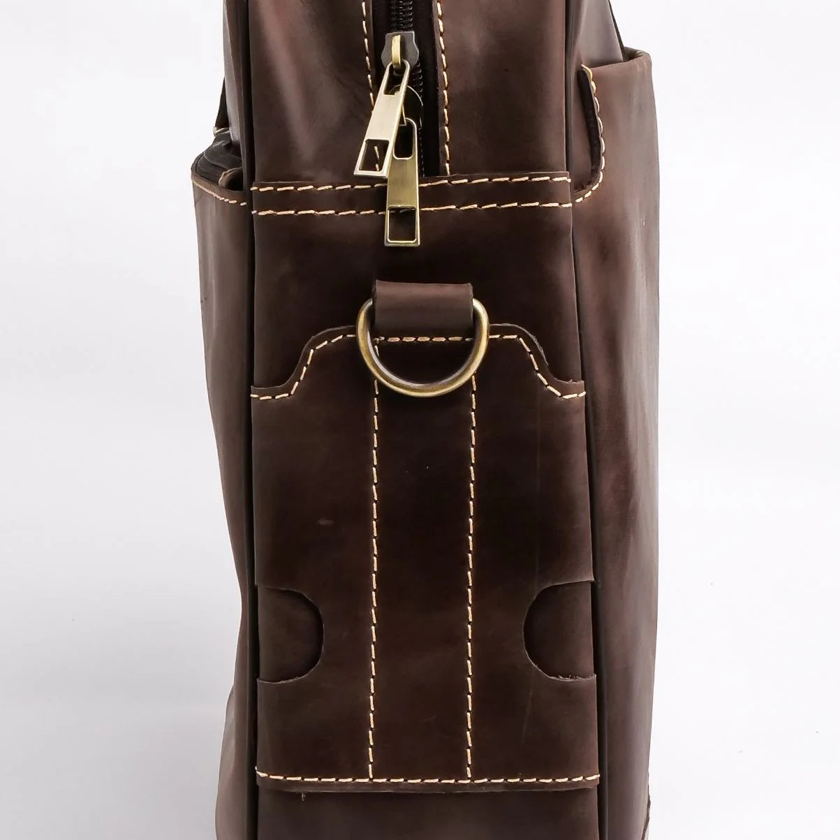Close-up of a dark brown leather bag showing a front pocket with contrast stitching, a brass D-ring, and a zipper with a gold-toned pull tab on a white background.