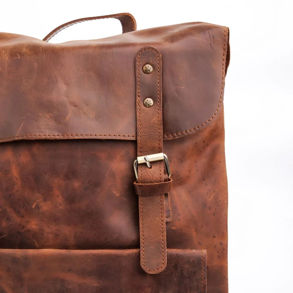 A close-up of a brown distressed leather backpack showing the front flap, a vertical strap with a metal buckle, and the texture of the leather against a white background.