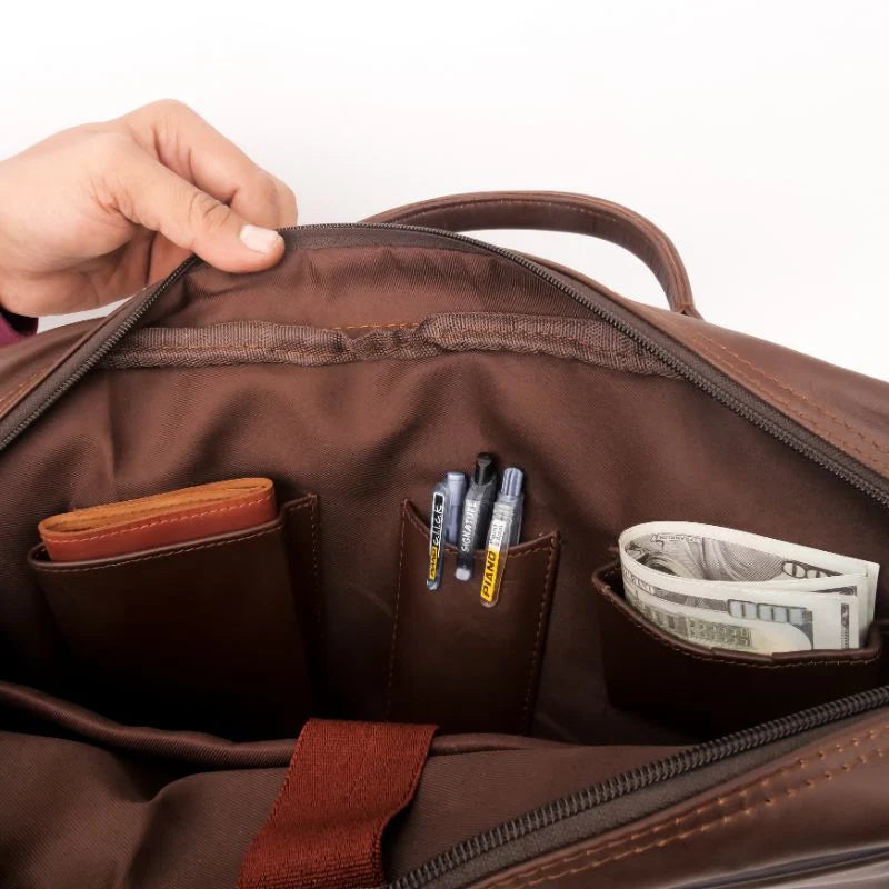 Open brown leather or faux-leather bag interior showing zip compartments and pockets holding a wallet, pens, and cash, with a hand holding the top edge.
